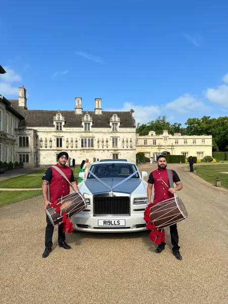 Two dhol players in red sherwanis beside a wedding car at a country house