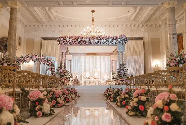 Floral mandap at the end of a long aisle lined with roses in a chandeliered hall