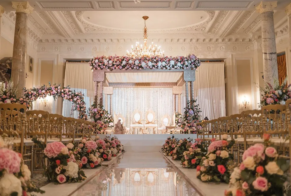 Floral mandap at the end of a long aisle lined with roses in a chandeliered hall