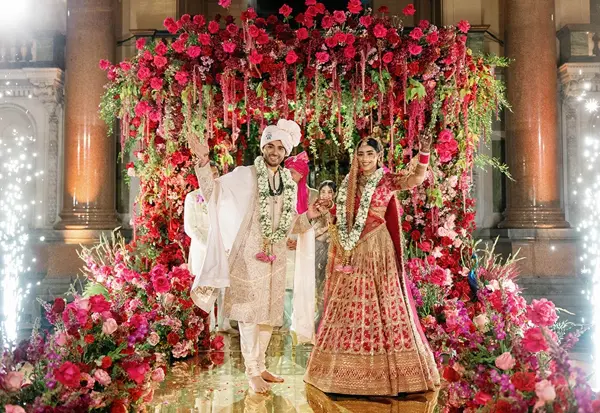 Bride and groom framed by a wall of red roses at the mandap