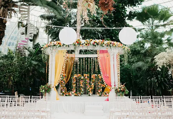 Mandap with lanterns and drapes in a palm-filled glasshouse