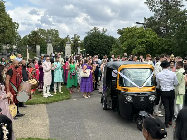 A tuk tuk leading a large wedding crowd with dhol