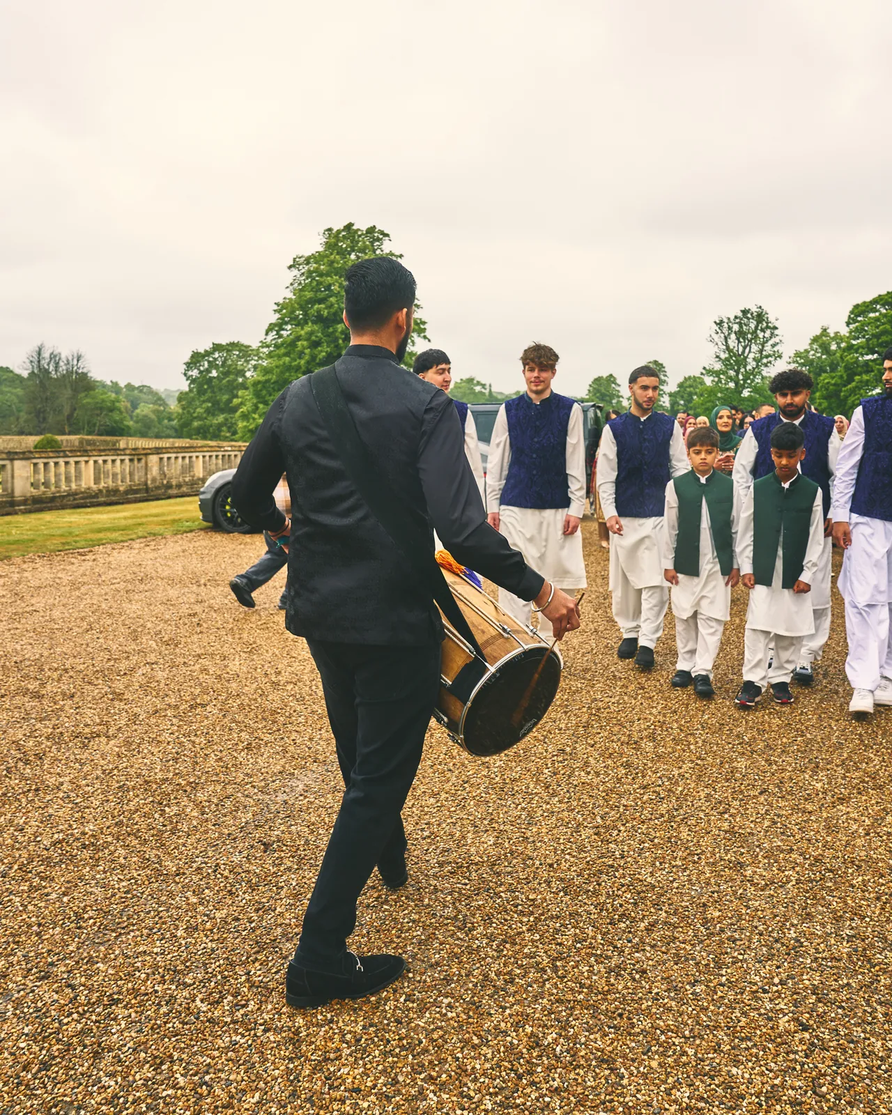 Dhol player leading the groom's party up a gravel drive