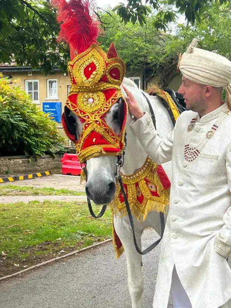 Groom in a cream sherwani beside a white horse in a red and gold headdress