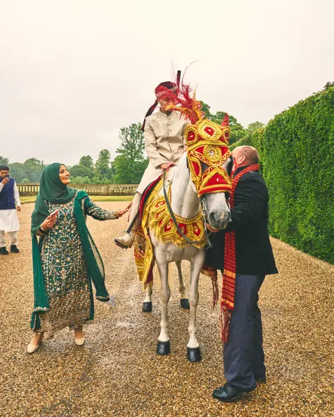 Groom on a white horse in red and gold baraat costume, welcomed by family