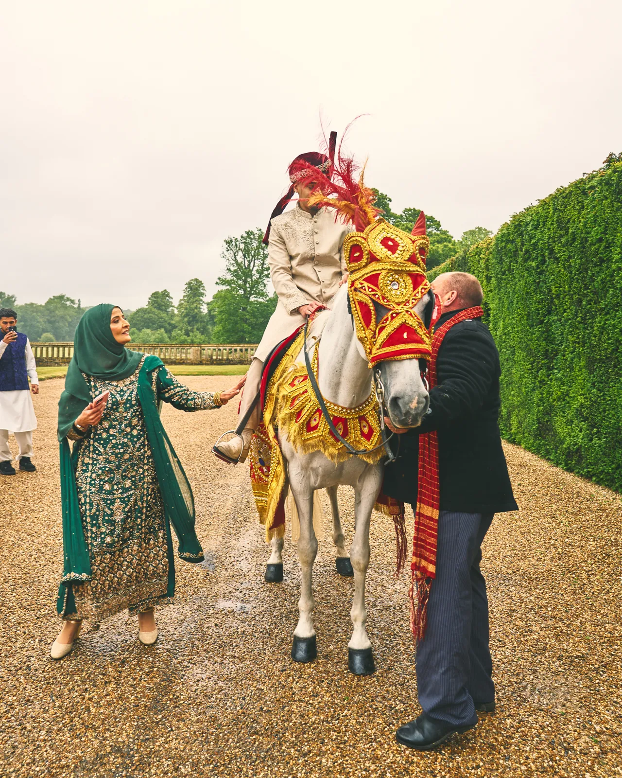 Groom on a white horse in red and gold baraat costume, welcomed by family